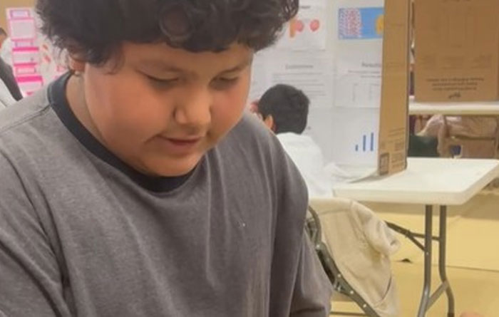 A student concentrates on a science fair project inside a classroom, with display boards and other students visible in the background.