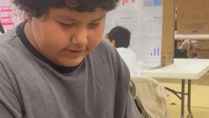A student concentrates on a science fair project inside a classroom, with display boards and other students visible in the background.